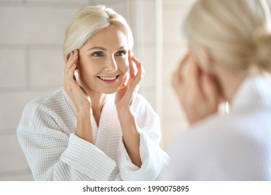 Gorgeous Mid Age Adult 50 Years Old Blonde Woman Standing In Bathroom After Shower Touching Face, Looking At Reflection In Mirror Smiling Doing Morning Beauty Routine. Older Dry Skin Care Concept.