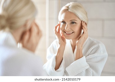 Gorgeous mid age adult 50 years old blonde woman standing in bathroom after shower touching face, looking at reflection in mirror smiling doing morning beauty routine. Older dry skin care concept.