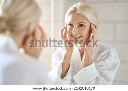 Gorgeous mid age adult 50 years old blonde woman standing in bathroom after shower touching face, looking at reflection in mirror smiling doing morning beauty routine. Older dry skin care concept.