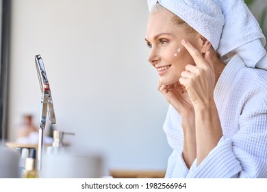 Gorgeous Mid Age Older Adult 50 Years Old Blonde Woman Wears Bathrobe In Bathroom Applying Nourishing Antiage Face Skin Care Cream Treatment, Looking At Mirror Doing Daily Morning Beauty Routine.