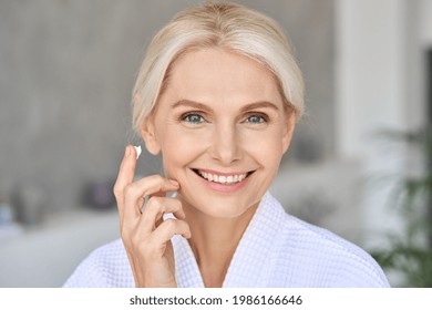 Gorgeous Mid Age Older Adult 50 Years Old Blonde Woman Wears Bathrobe In Bathroom Applying Moisturizing Sunscreen Face Skin Care Cream Treatment, Looking At Camera Doing Daily Beauty Routine.