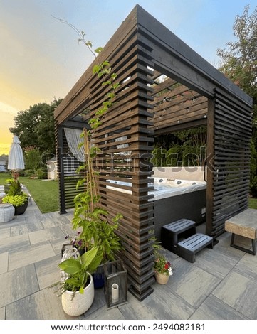 Gorgeous modern boho backyard design showcasing an outdoor pergola made from pine at sunset. Inside sits a hot tub spa with seats, lounger. Artsy decor and plant sets the mood with outdoor LED lights.