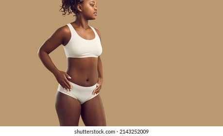 Gorgeous Plus Size Female Model Posing In Underwear. Confident Young African American Woman Wearing White Bralette And Underpants Standing With Hands On Hips On Solid Beige Color Text Space Background