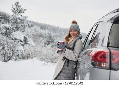 Gorgeous portrait. Pretty girl in warm clothes standing in the winter wood while leans on the car and holding cup of coffee.