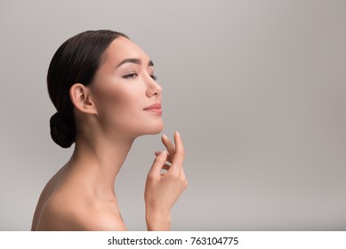 Gorgeous positive naked young asian woman with flawless skin is touching her face with pleasure while looking forward with slight smile. Isolated background and copy space in right side