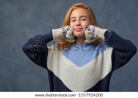 Gorgeous pretty young woman touches cheeks, pouts lips, has cheerful gaze. Teenage girl with long thick blonde hair wearing warm blue sweater and wool mittens isolated texture wall in Studio.