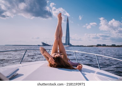 Gorgeous red haired woman laying on yacht with legs raised against background of the sea and modern skyscraper. Attractive Swedish woman on vacation at sea enjoying careless life. Rest and travel.