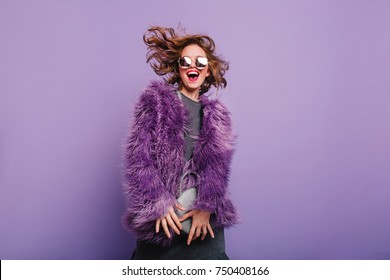 Gorgeous short-haired girl in sunglasses dancing on purple background with happy smile. Laughing female model in elegant fur coat posing in studio with little handbag.