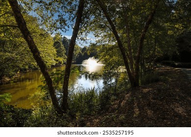 a gorgeous shot of a majestic water fountain in the middle of still lake water with lush green and autumn colored trees and plants reflecting off the lake water with blue sky and clouds at Lenox Park	 - Powered by Shutterstock