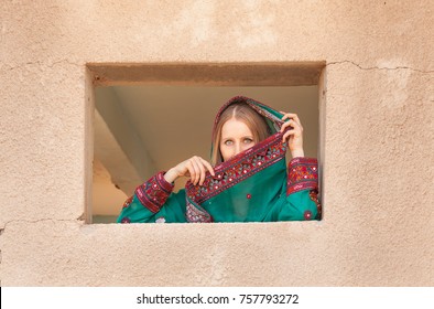 Gorgeous Shy Girl In Traditional Omani Clothes Looking Through The Window And Covering Her Face With Veil To Hide From Eyes Of People Walking Nearby 