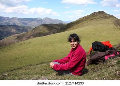 Gorgeous smiling young woman in purple sweater sitting on top of the grass covered mountain with her backpack to the side on a sunny morning with blue sky and clouds with space to the left - Powered by Shutterstock