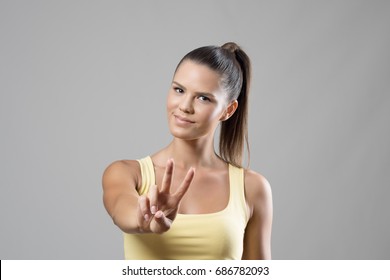 Gorgeous sporty young female portrait showing two finger peace hand sign over gray studio background. 