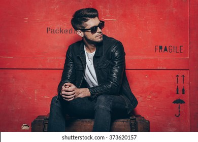 Великолепный и стильный. Красивый молодой человек в солнцезащитных очках держит руки сложенными и смотрит в сторону, сидя на деревянном сундуке на красном фоне