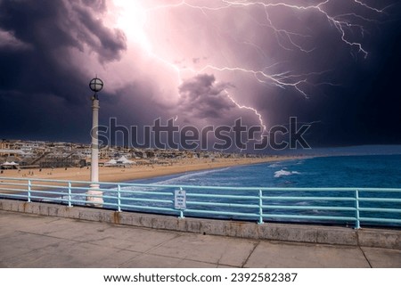 a gorgeous summer landscape at Manhattan Beach Pier with a blue metal hand rail and tall light posts along the pier, blue ocean water and people relaxing on the beach with storm clouds and lightning	