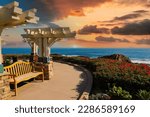 a gorgeous summer landscape at Treasure Island Beach with a footpath surrounded by lush green palm trees, grass and plants, blue ocean water and a white wooden pergola in Laguna Beach California USA