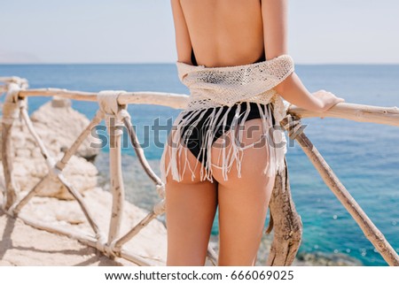 Gorgeous tanned lady in black swimwear standing near the wooden fence and looking at the ocean. Adorable young woman wearing trendy white knitted enjoying vacation at sea resort