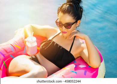Gorgeous tanned millennial young woman with sunglasses and jar with cold drink sitting in inflatable pink donut float in pool on sunny summer day. Vibrant colors. Retouched.