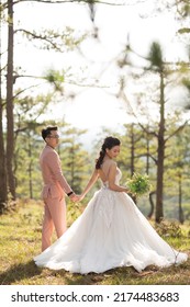 Gorgeous Wedding Couple In Mountain Of Green Pine Forest On Background. The Groom With In A Pink Shirt And The Bride In A Beautiful Dress On A Wedding Walk In The Woods In The Mountains.