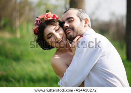 Gorgeous wedding portrait. Young handsome groom with beard in white shirt hugs his beautiful brunette bride with flowers in her hair. Sincere feelings and emotions. Tender wedding closeup. 