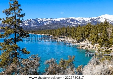 a gorgeous winter landscape with clear blue lake water, snow capped mountain ranges and lush green trees along the banks of the lake and blue sky at Lake Tahoe at Nevada State Park in Incline Village	