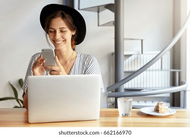 Gorgeous Woman In Black Hat And Blouse Smiling While Using Cell Phone Chatting With Her Boyfriend Sitting At Table With Laptop, Glass Of Water And Cake Enjoying Good Day. Beauty And Youth Concept