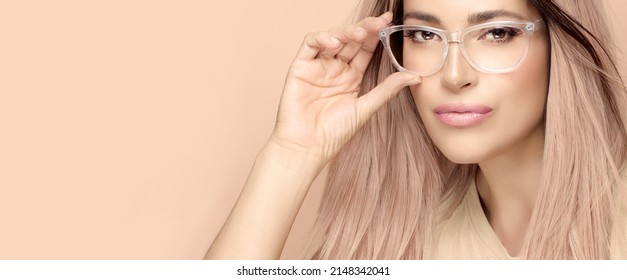 Gorgeous Woman Face With Trendy Clear Frame Glasses Posing In Studio. Beautiful Woman With Nude Makeup Adjusting Glasses Against Beige Background. Fashion Eyewear And Clear Vision Concept