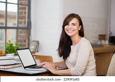 Gorgeous Woman With A Happy Beaming Smile Sitting At A Table Working At A Laptop Computer In Front Of A Window