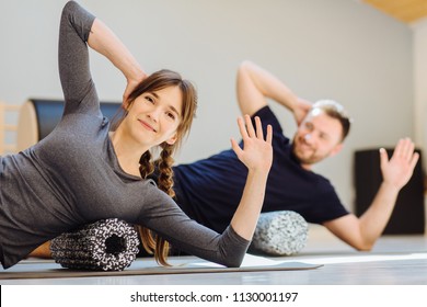 Gorgeous woman instructor performing back exercise on a foam roller at same time with handsome guy at pilates studio. Coach and patient doing fascia exercise on side surface of back.
