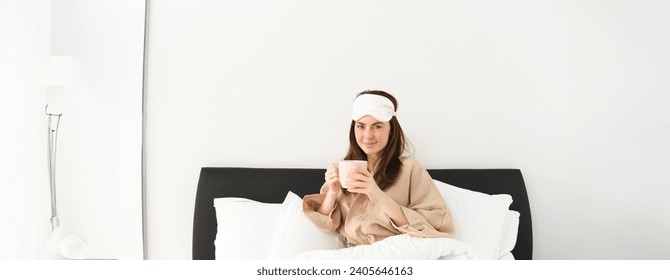 Gorgeous woman in pyjamas and sleeping mask, holding cup of coffee, drinking cappuccino in her bed and looking happy, resting in bedroom on weekend, having relaxing morning.