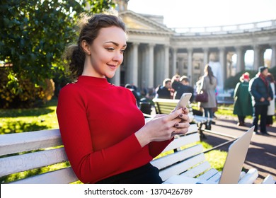 Gorgeous Woman University Student Reading Article On Site Via Mobile Phone During Online Learning Via Notebook Device, Sitting On A Campus In Spring Day. Pretty Female Using Apps On Cell Telephone