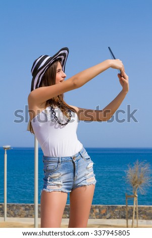 Gorgeous young busty naughty long haired brunette teen girl in jean shorts and hat, enjoying the early summer morning sun by the ocean, posing for selfies using a tablet.