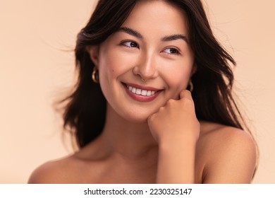 Gorgeous young delighted Asian female model with long dark hair and perfect skin touching cheek smiling and looking away against beige background