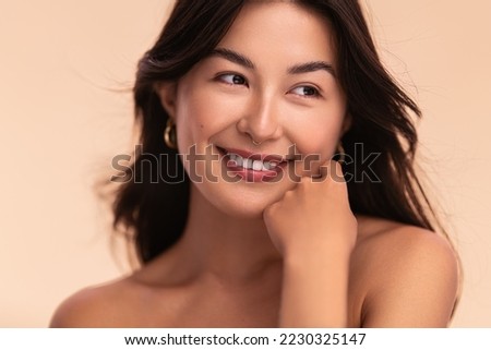 Gorgeous young delighted Asian female model with long dark hair and perfect skin touching cheek smiling and looking away against beige background