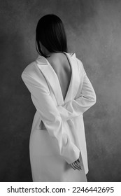 Gorgeous young female in classic white jacket. Brunette with beautiful back. Grey background. Photo Studio. Stylish. Black and white. Soft light. Man clothes. Fashion editorial. Elegant and sexy.