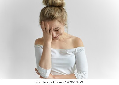 Gorgeous young female wearing off shoulder white top keeping hand on her face, covering one half, feeling shy. Charming cute girl with hair knot looking down and laughing at awkward moment