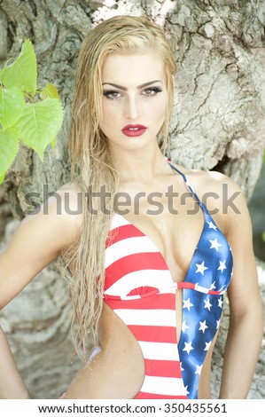 A gorgeous young fit and petite blonde with wet hair looking at the camera wearing an American flag one piece swimsuit and a tree in the background on a sunny day.