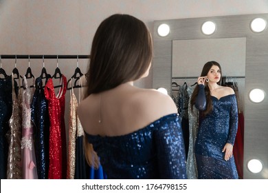 Gorgeous young lady in luxury outfit standing in front of the mirror trying on earings. Selective focus on the mirror. Luxury gowns on hangers in the rent shop for ball, wedding, or photoshot.