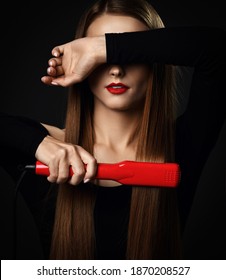 Gorgeous young slim woman with long silky straight hair in black bodysuit uses red hair straightener and covers eyes with her arm over dark background. Haircare, beauty, wellness concept