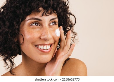 Gorgeous young woman looking away with a smile while applying moisturising cream on her face. Happy young woman treating her facial skin with a nourishing beauty product. - Powered by Shutterstock