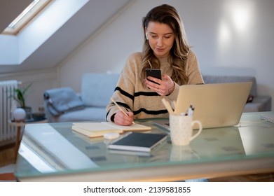 Gorgeous Young Woman Sitting At Desk Behind Her Laptop And Text Messaging On Her Smartphone While Working From Home. Home Office.