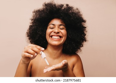 Gorgeous Young Woman Smiling Cheerfully While Dropping Cosmetic Face Serum Onto Her Palm. Happy Woman With Afro Hair Treating Her Skin With A Moisturizing Beauty Product.