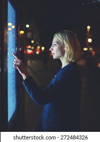 Gorgeous Young Woman Touching Big Digital Screen While Consults About Touristic Information In The Night City With Beautiful Urban Lights On Background, Female Use Modern Kiosk Looking To Blue Display