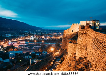 Gori, Shida Kartli Region, Georgia. Walls Of Gori Fortress And Cityscape In Evening Illumination Under Blue Sky. Travel Destination In Night Lights. Goris Tsikhe Is A Medieval Citadel