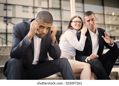 Gossip Colleagues In Front Of Their Office Sitting On Stairs, Depressed Businessman Portrait And Gossip Out Of Focus In Background. Reason Is Racism. Shallow Depth Of Field.