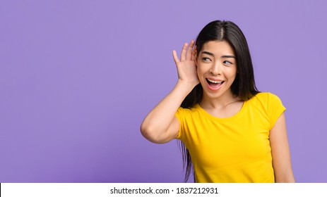 Gossip Concept. Curious young asian woman holding hand near ear, trying to hear interesting information, cheerful korean female eavesdropping on purple studio background, panorama with copy space