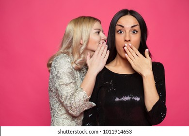Gossip. Two young beautiful smiling girlfriends in little black dresses. The blonde tells a brunette story or gossip in her ear. - Shutterstock ID 1011914653