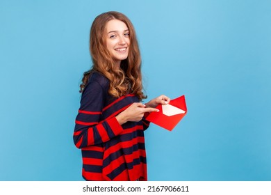 I got love letter on Valentine's day. Woman in striped casual style sweater, pointing letter in red envelope, holding greeting card and smiling joyfully.Indoor studio shot isolated on blue background. - Shutterstock ID 2167906611