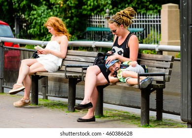 Goteborg, Sweden - July 25, 2016: Young Adult Woman Using Phone While Patting A Boy On His Head As He Rest It On Her Lap. Woman In Background Reading A Book. Real People In Everyday City Life.