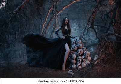 A gothic beautiful young woman in a black dress stands near a mountain of human skulls. Fashion model in the image of the goddess of death in a dark forest of trees. Silk fabric flies in the wind.