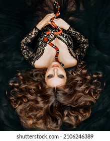 Gothic fantasy woman dark queen of snakes and poisons. Fairy girl princess lies, hugs red snake sinaloa. Long flowing hair, black vintage medieval lace dress. Royal milk snake crawls on chest and face